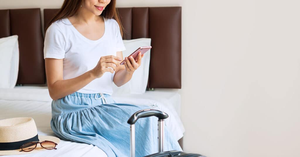 Woman sit on a bed of a hotel room using a mobile device with hospitality solutions for guests