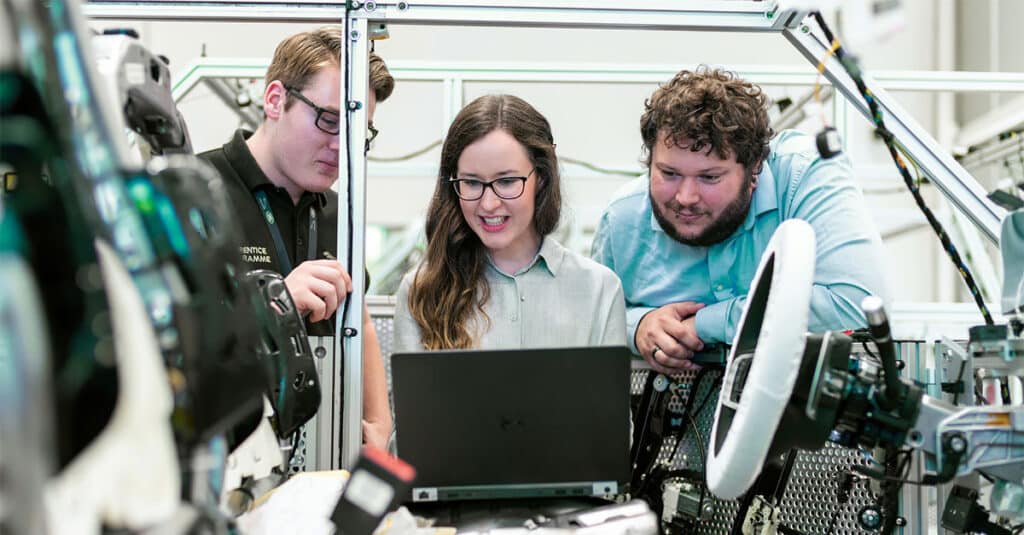 Three automotive engineers collaborating on a laptop connected to a prototype car, showcasing automotive industry transformation