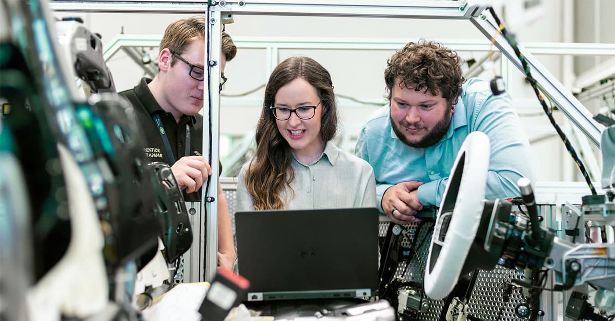 Three automotive engineers collaborating on a laptop connected to a prototype car, showcasing automotive industry transformation