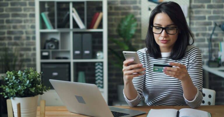 A woman sits in front of a laptop, holding a phone and a credit card, suggesting an unsafe online transaction.
