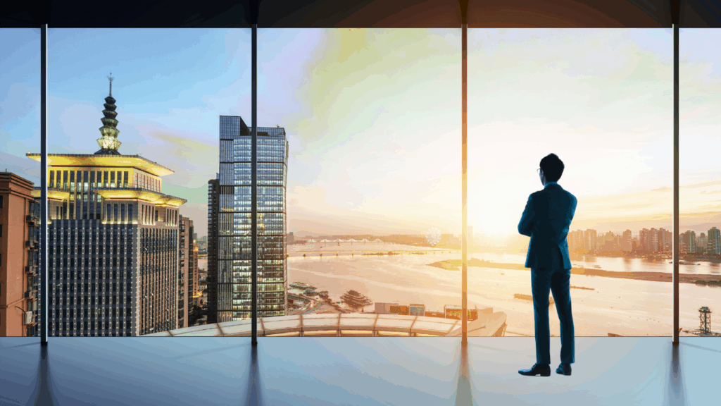 Silueta de un hombre de negocios vestido con traje de pie junto a la ventana de una oficina que va del suelo al techo, contemplando el horizonte de la ciudad al amanecer.